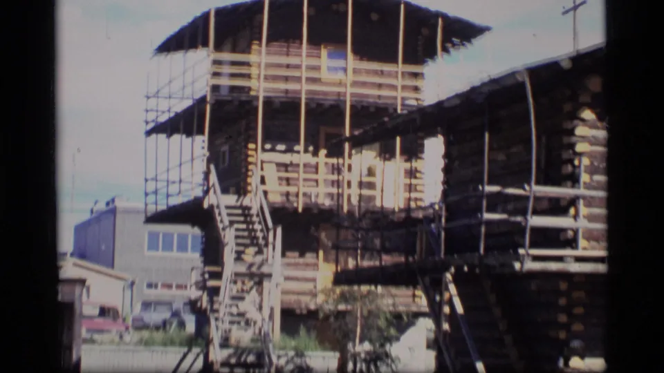 1977 Alaska 8mm frame showing the log cabin from another angle, with scaffolding, exterior stairs, railings, and an open upper level.