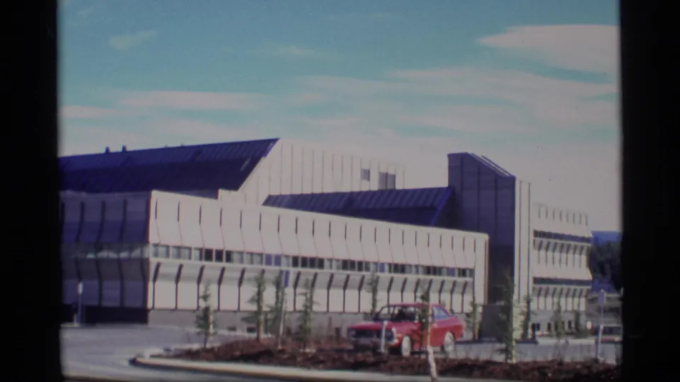 1977 Alaska 8mm frame of a modern white building with dark roof panels, paved frontage, young trees, and a red car.
