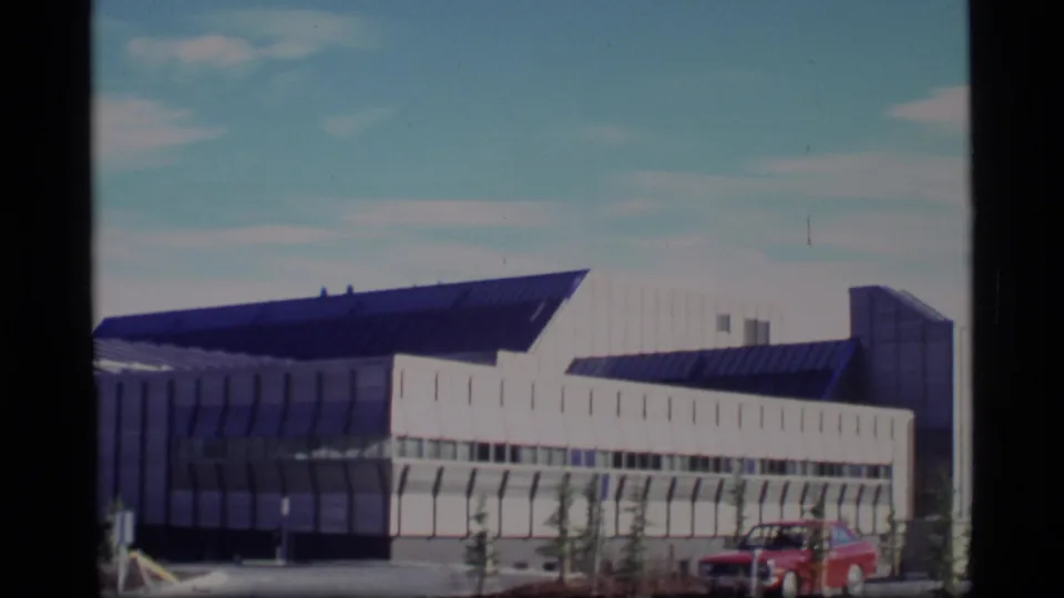1977 Alaska 8mm frame showing the side of a modern building with repeating wall panels and a red car near the curb.
