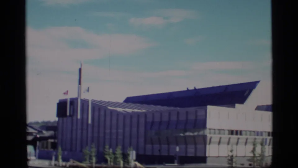 1977 Alaska 8mm frame showing a modern building corner, flagpoles, young trees, and a low dark roofline.