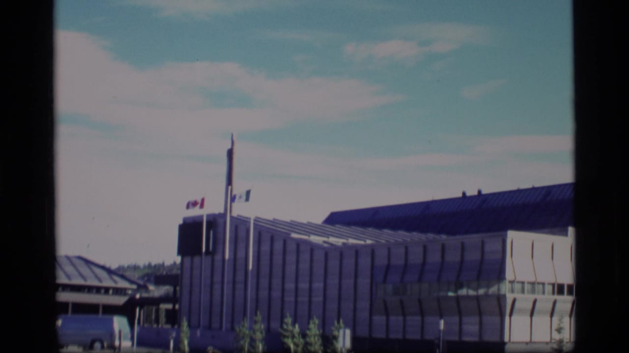 1977 8mm frame showing a modern civic-looking building with Canadian and blue-and-white flags near a tall pole.