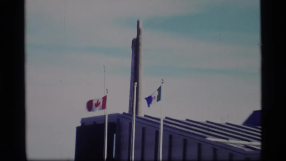 Closer 1977 8mm view of Canadian and blue-and-white flags flying beside a tall Government of Yukon pole.