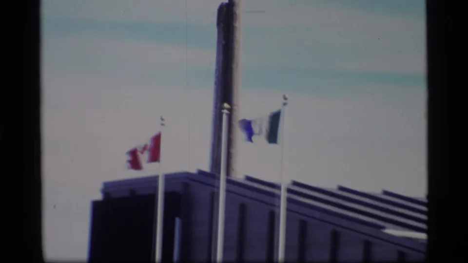 1977 home-movie frame with Canadian and blue-and-white flags beside a tall pole over a modern civic roofline.