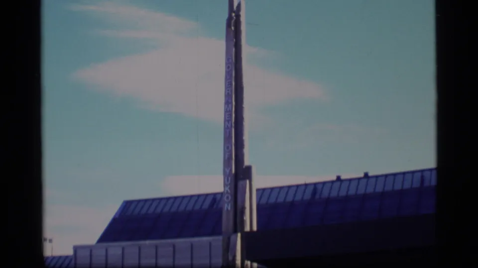 1977 8mm frame showing the upper facade of a glass-roofed building with a tall Government of Yukon sign against the sky.