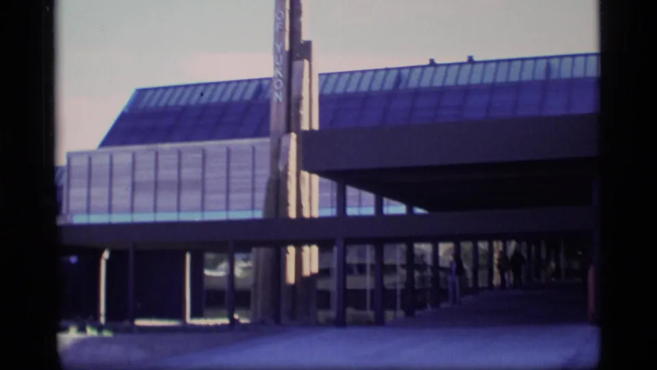 1977 home-movie frame of a public building entrance with glass roof panels, vertical Government of Yukon sign, and a covered walkway.