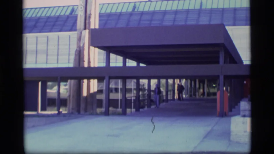 1977 8mm frame showing the Government of Yukon building exterior, covered walkway, pavement, and small pedestrians near the entrance.