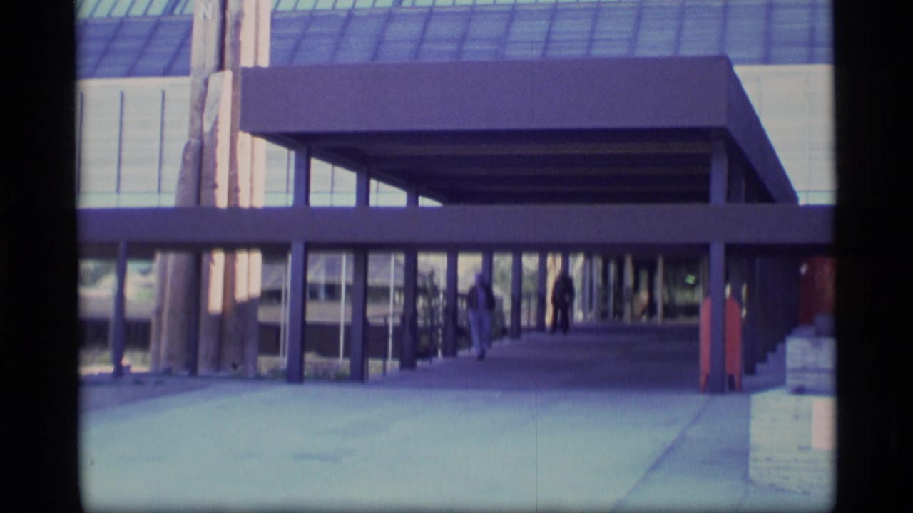 1977 Alaska 8mm frame showing a glass-fronted modernist building, covered walkway, and distant visitors near the entrance.