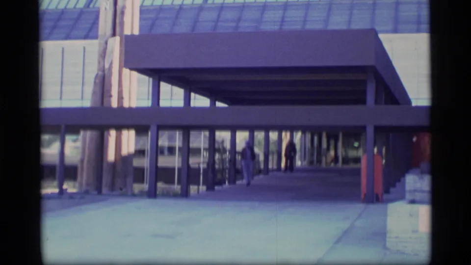 1977 Alaska 8mm frame showing a glass-fronted modernist building, covered walkway, and distant visitors near the entrance.