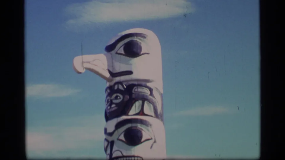 1977 Alaska 8mm frame showing the upper carved figures of a totem pole set against a pale blue sky.