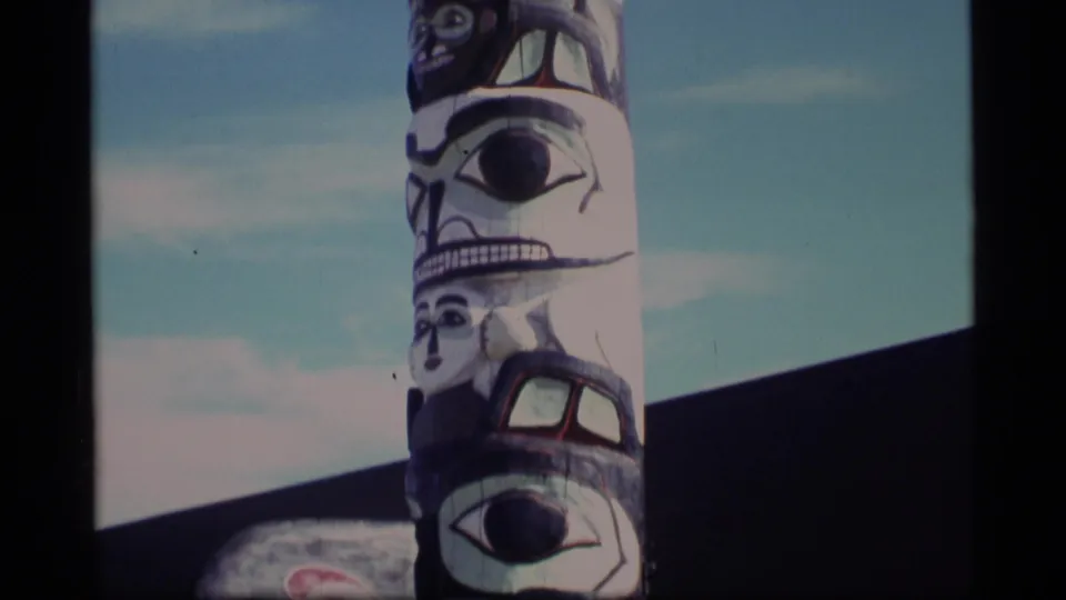 1977 Alaska 8mm frame showing more of the totem pole beside a dark building roofline and open sky.