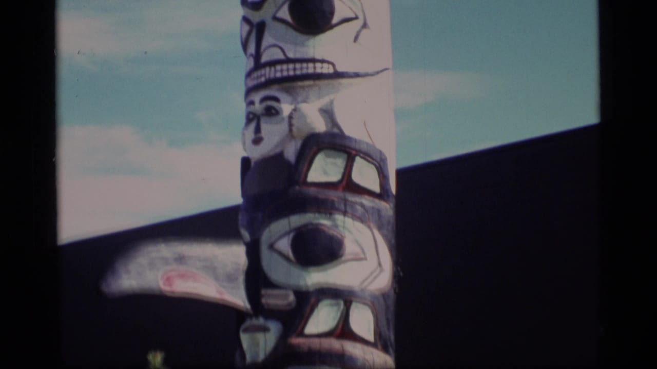 1977 Alaska 8mm frame showing the upper portion of a painted totem pole with carved faces, set against blue sky and a dark wall.