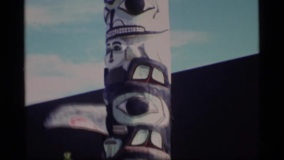 1977 Alaska 8mm frame showing the upper portion of a painted totem pole with carved faces, set against blue sky and a dark wall.