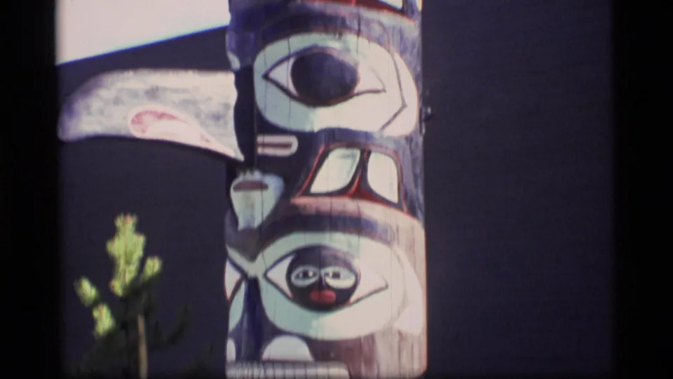Close 8mm view of a painted totem pole in Alaska, with stacked carved faces and black, white, red, and pale green painted areas.