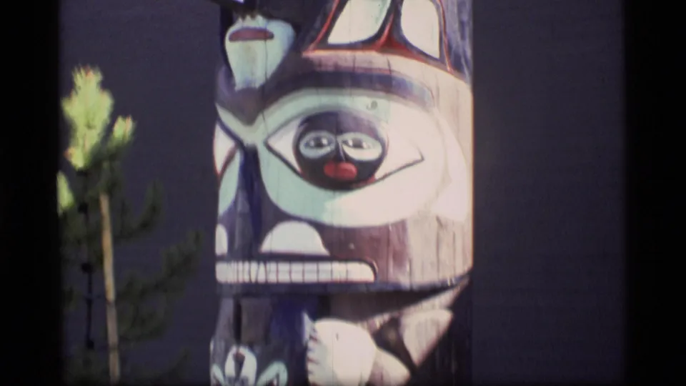 1977 Alaska home-movie frame showing the lower middle of a painted totem pole, partly obscured by a small evergreen in the foreground.