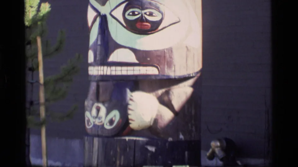 Alaska 8mm frame from 1977 showing the lower portion of a painted totem pole, including a carved face, hand-like forms, and a small tree nearby.