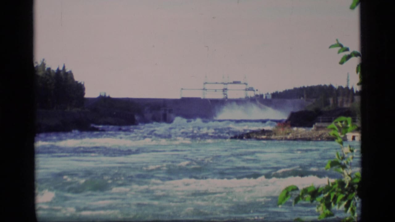 1977 Alaska 8mm frame showing turbulent water below a hydroelectric dam with forested banks and foreground foliage.