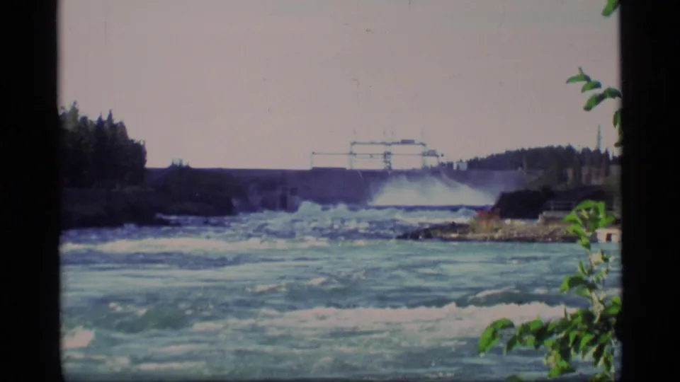1977 Alaska 8mm frame showing turbulent water below a hydroelectric dam with forested banks and foreground foliage.