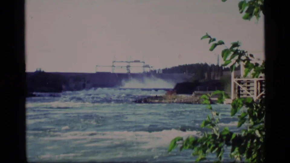 1977 Alaska 8mm frame of blue-green rapids below a dam, with foreground leaves and a small riverside structure.