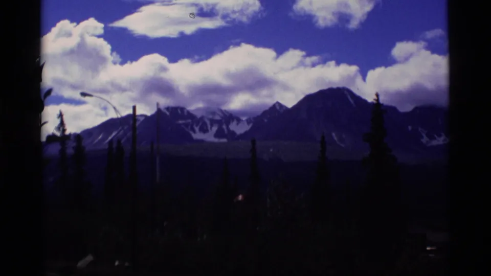 1977 Alaska travel footage frame showing snow-streaked mountains, dramatic clouds, and dark evergreen silhouettes.