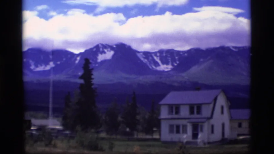 1977 Alaska 8mm frame showing a white two-story rural house below snow-streaked mountains, with trees and open ground nearby.
