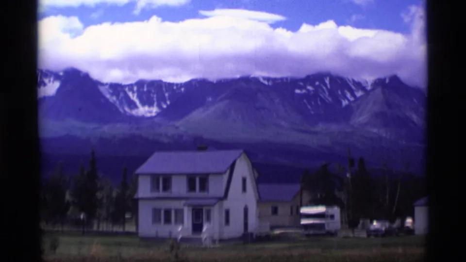 1977 Alaska 8mm frame of a white rural house, parked vehicles, trees, and snow-streaked mountains under a bright cloud bank.
