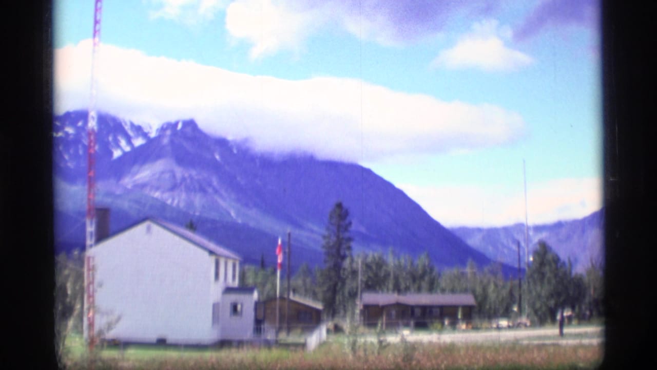 1977 Alaska home-movie frame with a white house, roadside buildings, trees, and mountains beneath large clouds.