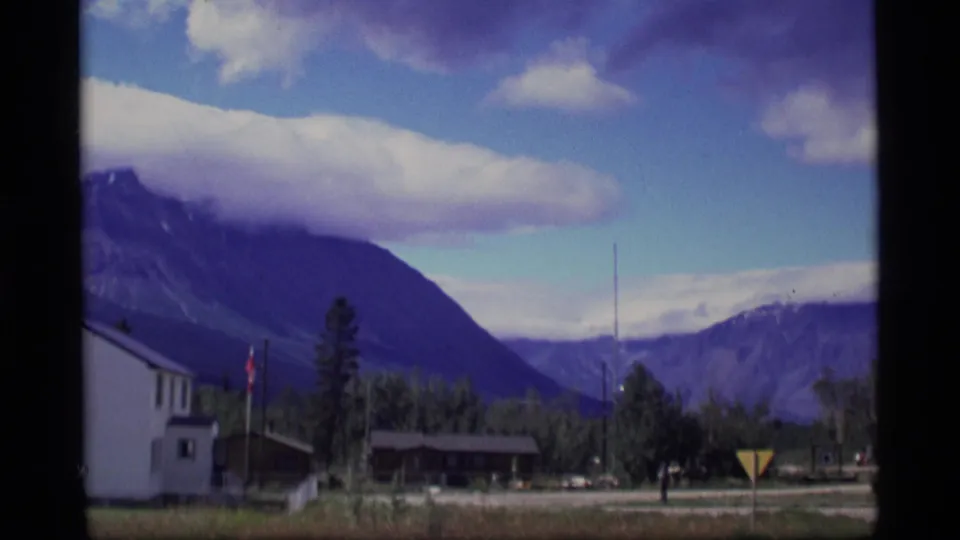 1977 Alaska frame showing roadside buildings, trees, utility poles, a triangular sign, and mountains in the distance.