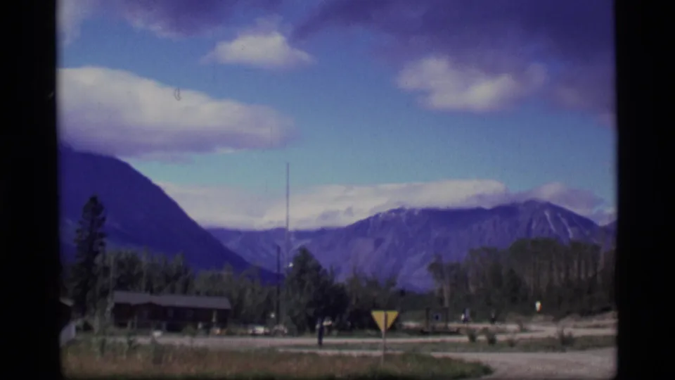 1977 Alaska home-movie view of a road, cabin, trees, mountains, a sign, and a distant person near the roadside.