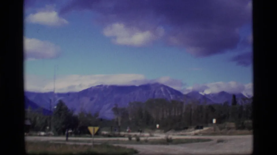 1977 Alaska frame with an open road, triangular sign, trees, distant figures, and mountains under a partly cloudy sky.