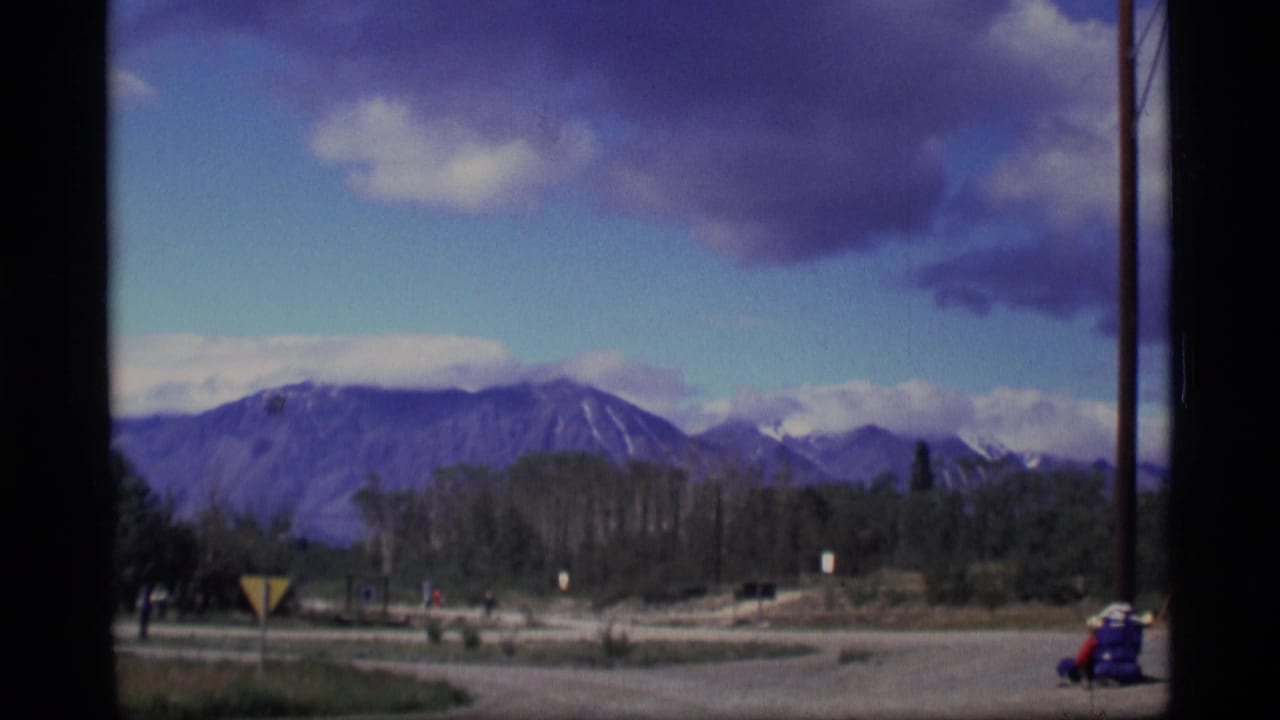 Wide roadside view with distant mountains, trees, road, yellow sign, and a small person in red near the edge.