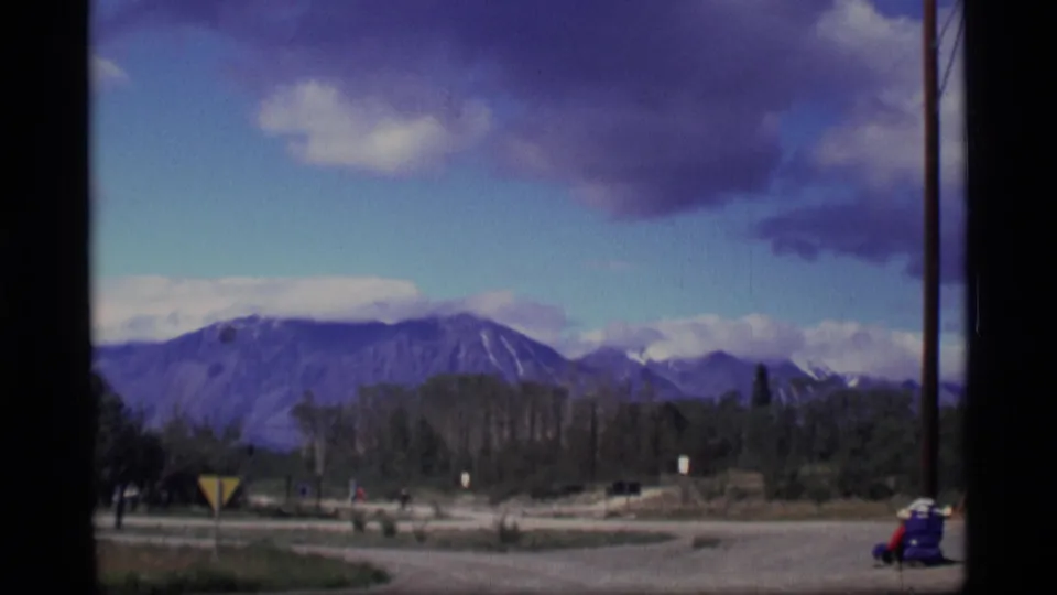 1977 Alaska 8mm frame showing distant mountains beyond trees and a road, with a yellow warning sign and a small person in red at right.