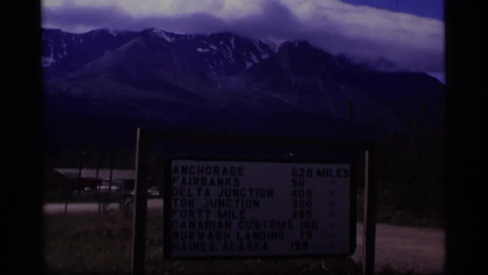 1977 Alaska roadside distance board in front of mountains and clouds, with a road and trees partly visible in low exposure.