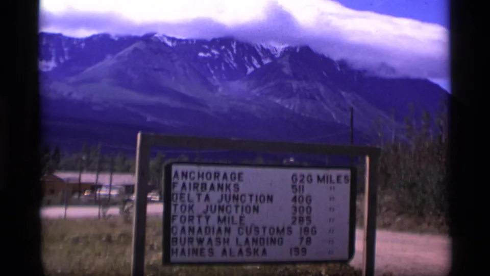 1977 Alaska 8mm view of a distance sign naming Anchorage, Fairbanks, Delta Junction, Tok Junction, and other destinations below mountains.