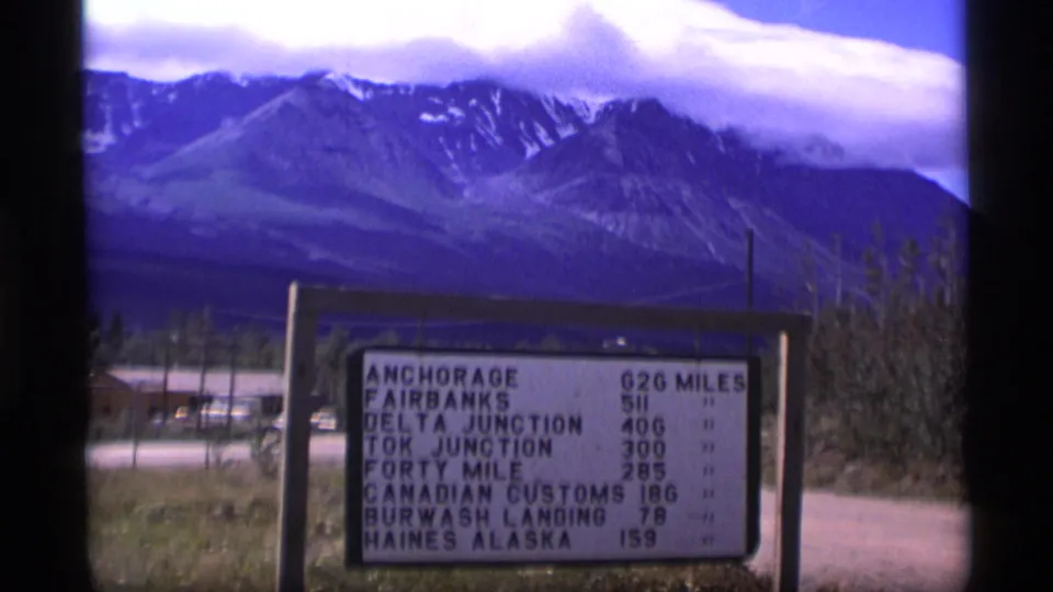 1977 Alaska frame showing a roadside mileage sign against snow-streaked mountains, with destination names and distances partly readable.