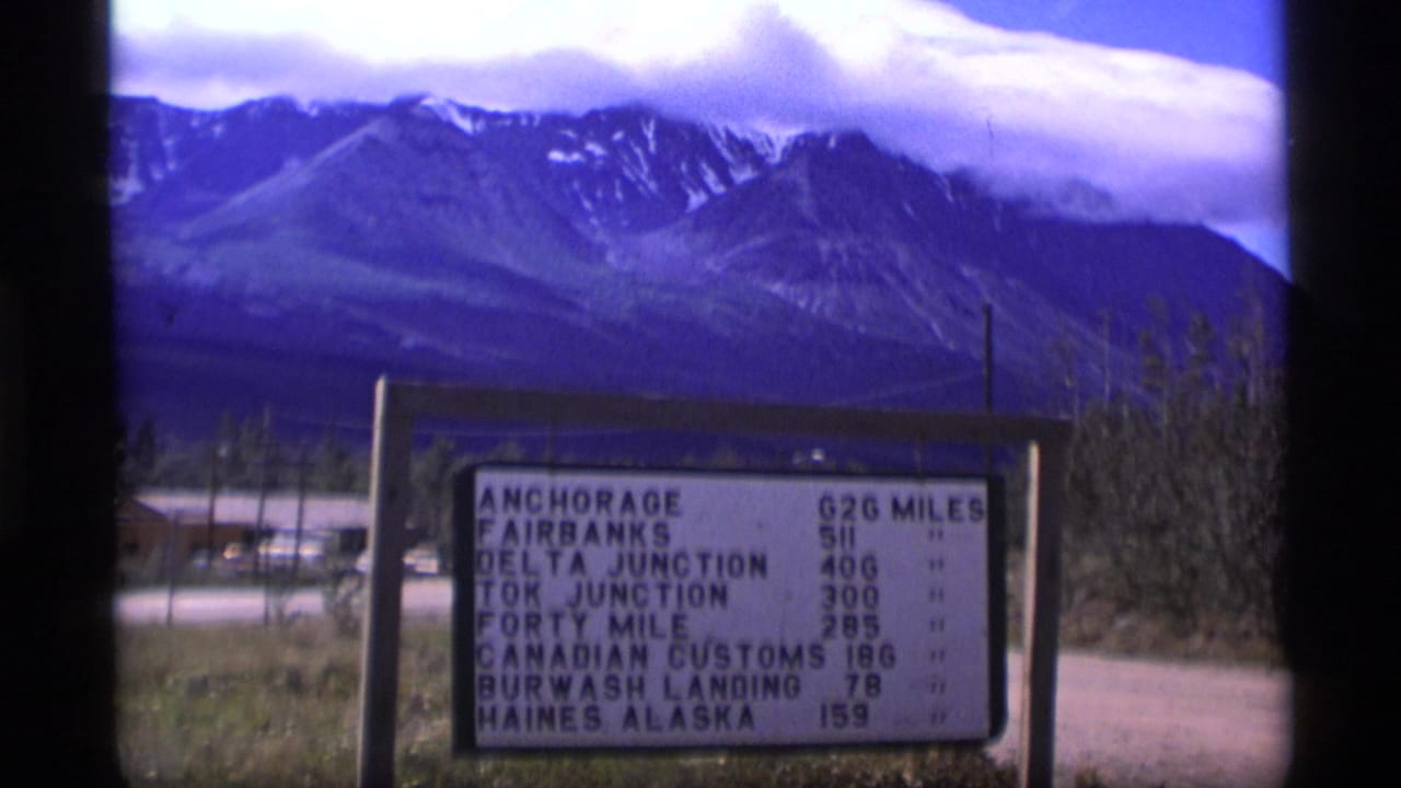 1977 Alaska home-movie frame showing a roadside mileage sign with mountains and low cloud behind it.