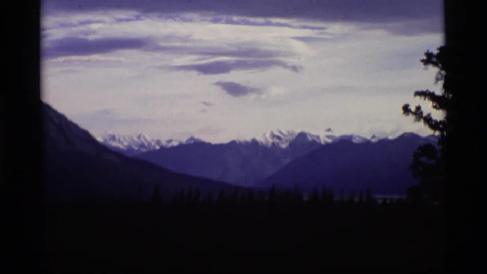 1977 Alaska Super 8 frame with distant snow-capped summits beyond a dark valley and evergreen silhouettes.