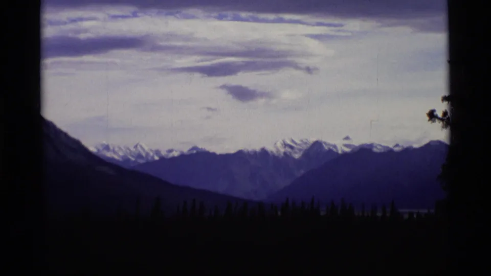 1977 Alaska home-movie landscape showing distant snowy ridges across a dark forested valley under a pale sky.