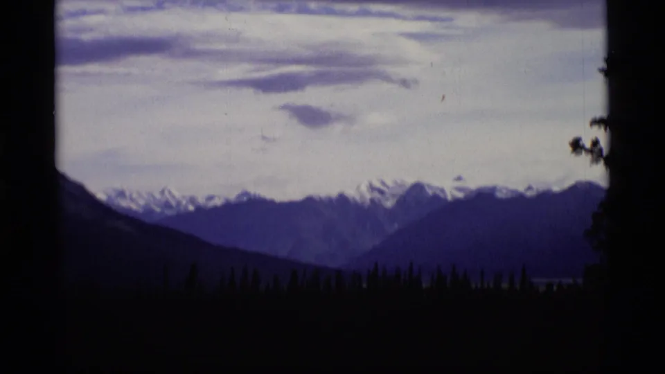 1977 Alaska 8mm frame showing shadowed evergreens and distant snow-capped mountains beneath a cloudy sky.