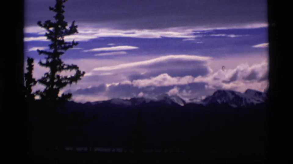 1977 Alaska 8mm frame with a tall evergreen silhouette, dark mountain ridge, snow peaks, and layered clouds.