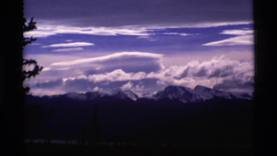 1977 Alaska 8mm frame showing a dark evergreen edge, distant snow-capped peaks, and dramatic layered cloud bands.
