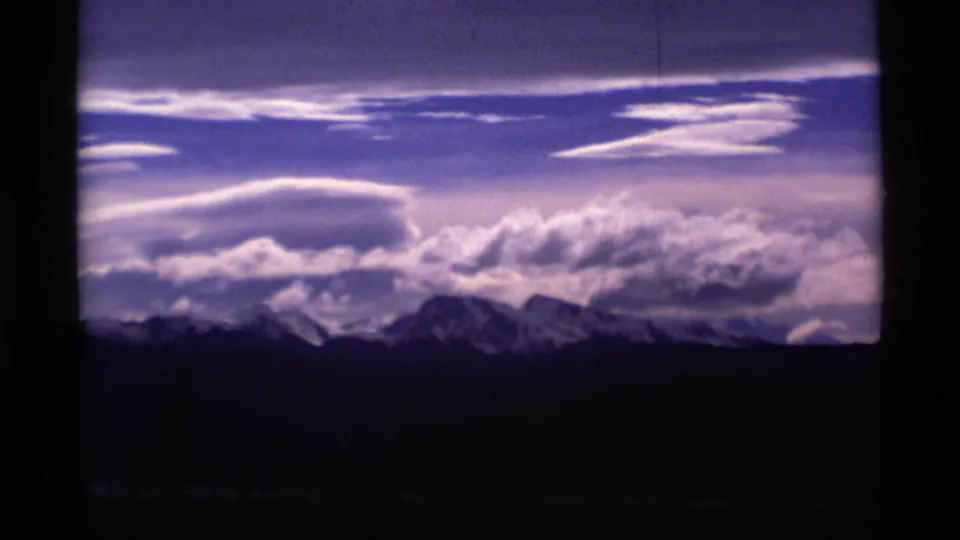 1977 Alaska 8mm frame with snow-capped mountains, sweeping white cloud layers, and a dark silhouetted foreground.