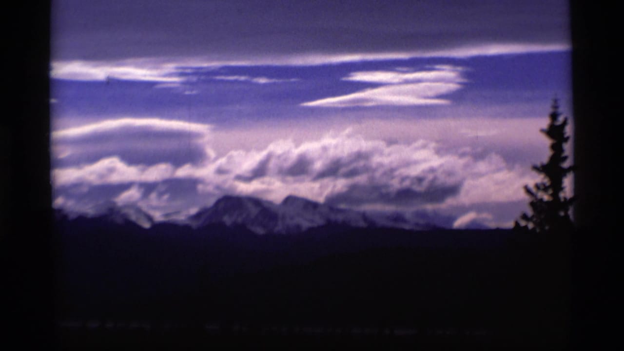 1977 Alaska 8mm frame showing distant snow-capped mountains, dramatic clouds, and a dark evergreen silhouette in the foreground.