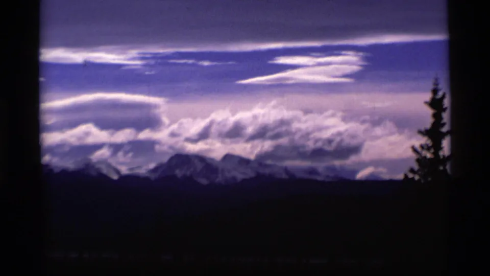 1977 Alaska 8mm frame showing distant snow-capped mountains, dramatic clouds, and a dark evergreen silhouette in the foreground.