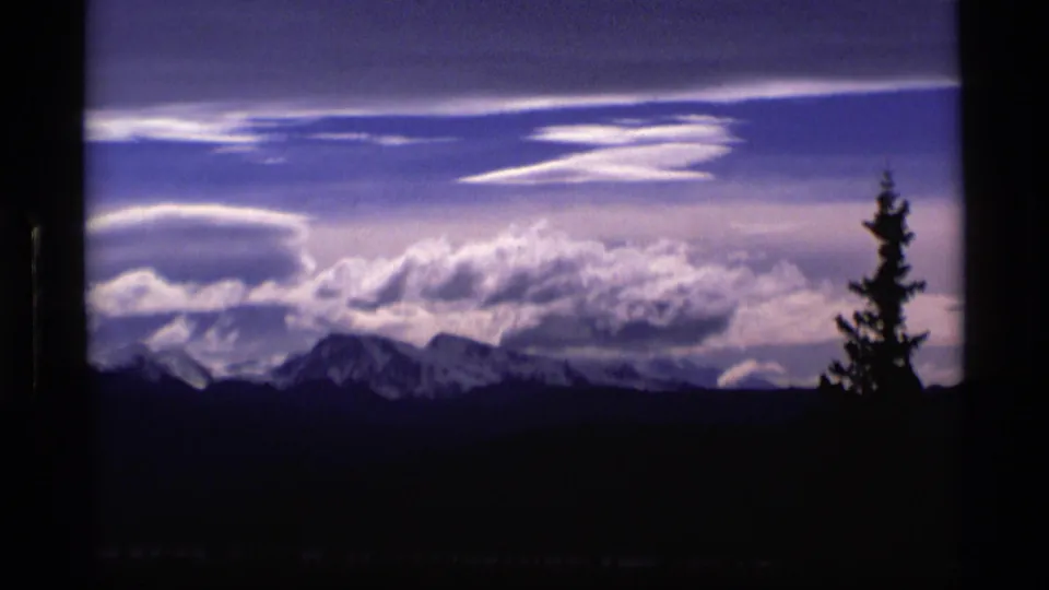 1977 Alaska home-movie frame with snow-capped peaks, broad cloud bands, and a dark conifer silhouette framing the right side.