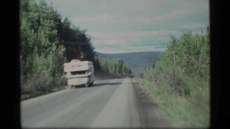1977 Alaska 8mm travel view showing a white RV on a forest road, with dense roadside vegetation and distant hills ahead.