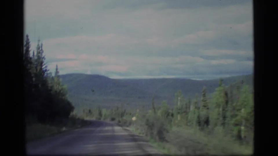 1977 Alaska home-movie frame showing a winding forest road, evergreen slopes, distant hills, and a pale overcast sky.