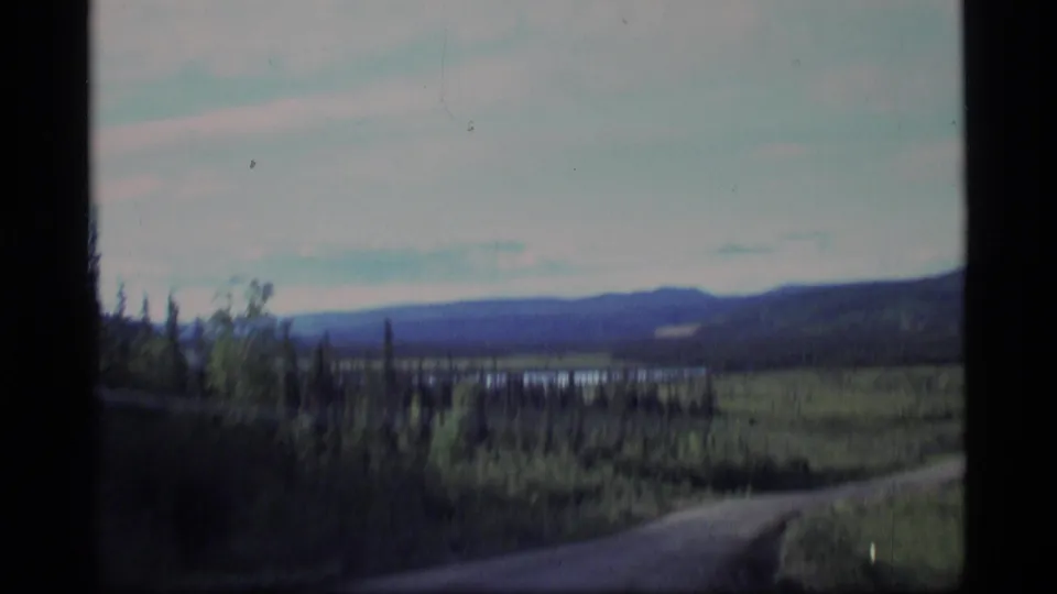 1977 Alaska home-movie frame from a moving vehicle, with a curved road, spruce trees, hills, and a possible lake.