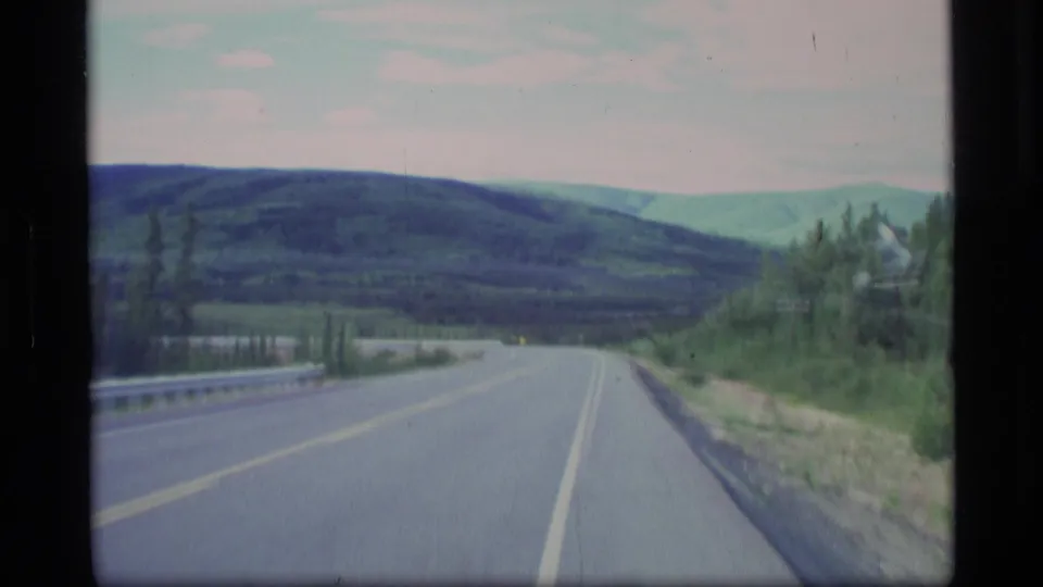 1977 Alaska 8mm frame showing a two-lane road, guardrail, roadside trees, and layered green mountain slopes.