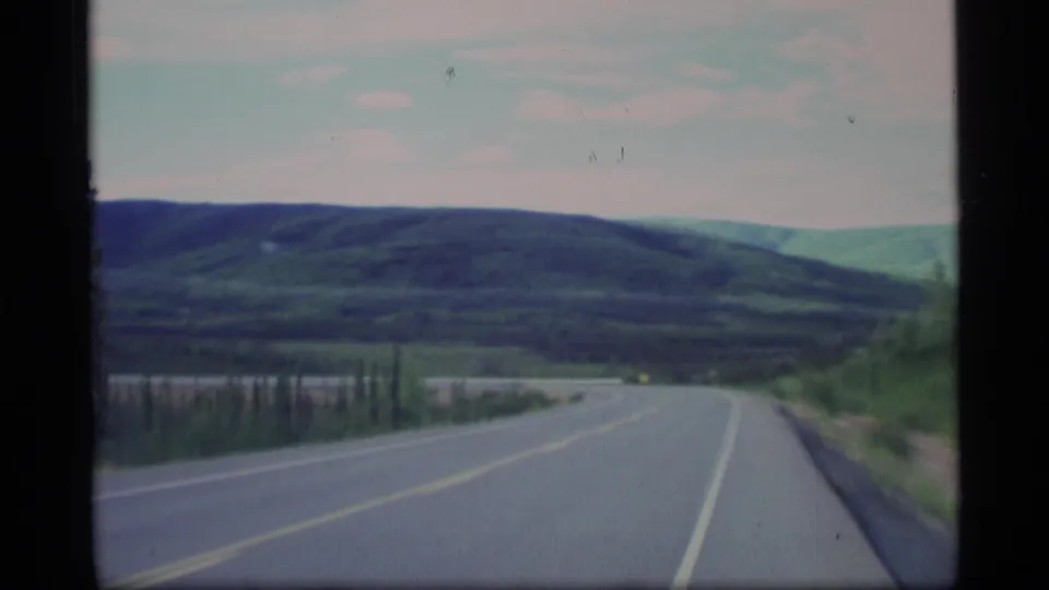 1977 Alaska road-trip footage showing a bend in the paved road beside trees, hills, and a pale overcast sky.