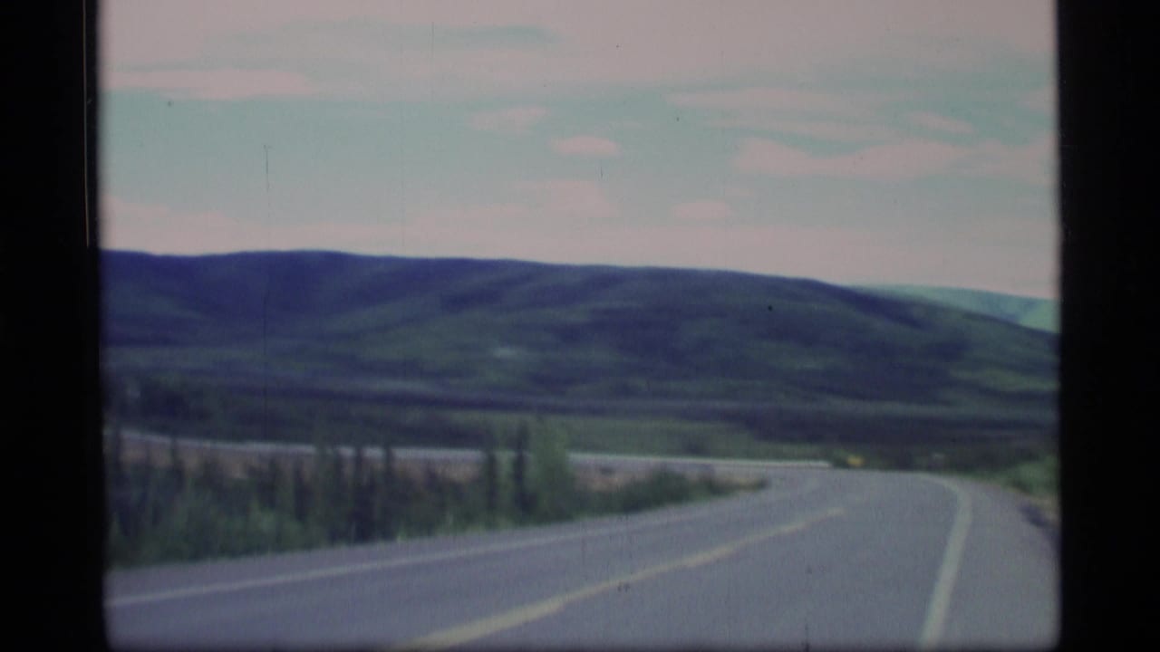 1977 Alaska 8mm frame showing a quiet two-lane road filmed from a moving vehicle, with green hills and trees ahead.